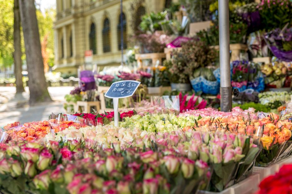 A vibrant outdoor flower market display featuring numerous bouquets of fresh roses and other flowers arranged on tables. The flowers include shades of pink, white, peach, and orange, with some wrapped