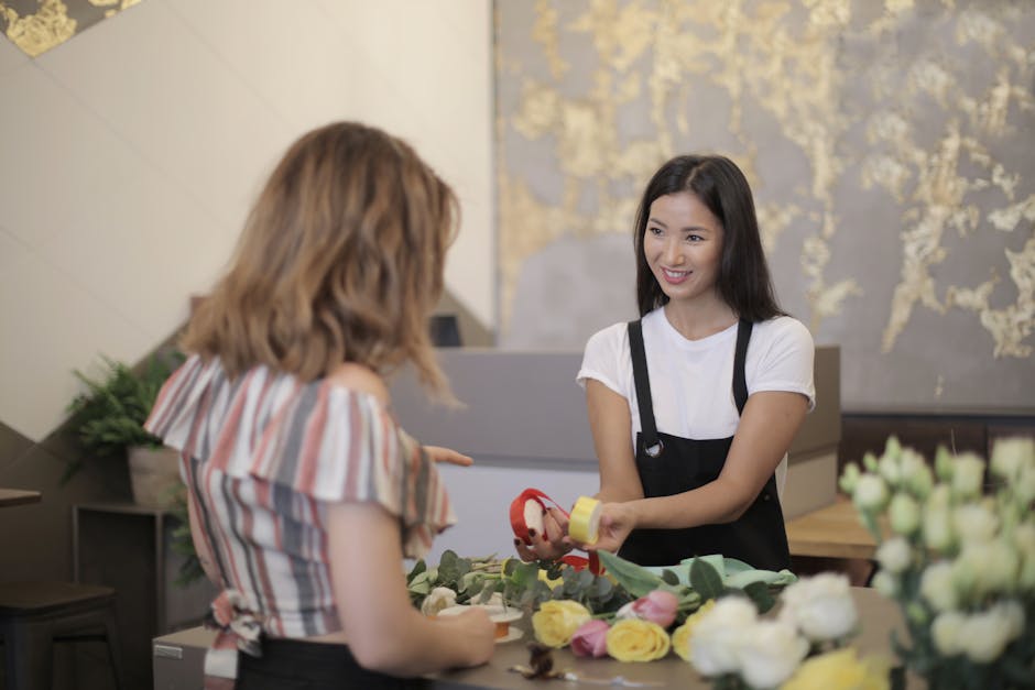 A young woman with long dark hair, wearing a white t-shirt and black apron, is smiling as she hands over a floral gift wrapped in yellow paper with a red ribbon to a customer in a flower shop. The cus