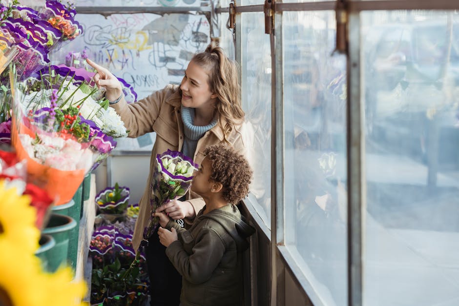 A woman and a young child at a flower shop display, with the woman smiling and pointing at a variety of vibrant flower bouquets arranged on shelves. The flowers include purple and white petunias, red,