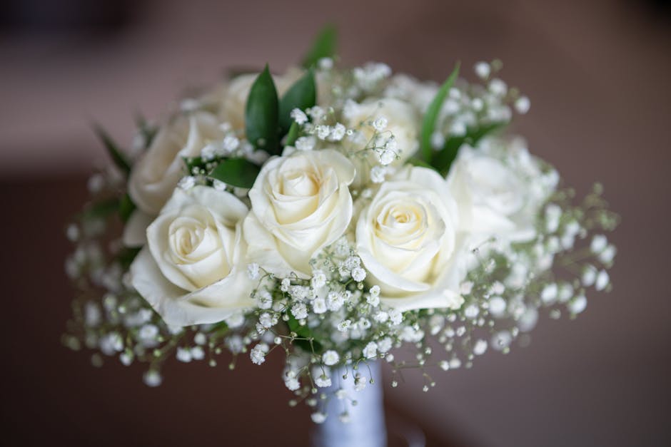 A bouquet of fresh ivory-white roses with tightly layered petals arranged in a rounded form, accented with delicate sprigs of baby's breath and dark green foliage, resting on a neutral surface. The ro