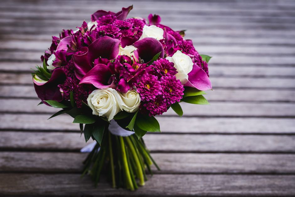 A bouquet of fresh flowers arranged with deep magenta calla lilies, bright pink chrysanthemums, and purple dendrobium orchids, accented by white roses and lush green leaves. The flowers are wrapped in