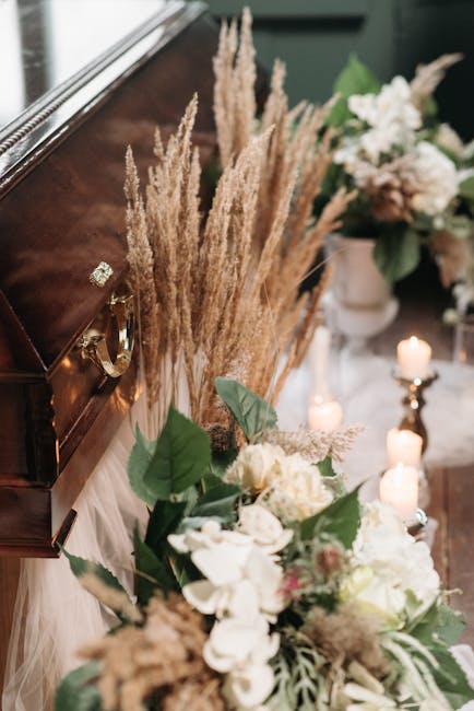 A floral arrangement in a softly lit indoor setting, featuring a bouquet of white and cream-colored flowers with green leaves placed near a dark wooden piece of furniture. Behind the bouquet, tall, de