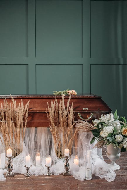 A floral arrangement featuring large white and pale blush roses, white peonies, and assorted greenery in a white vase, placed on a wooden table decorated with white tulle fabric. Surrounding the vase 